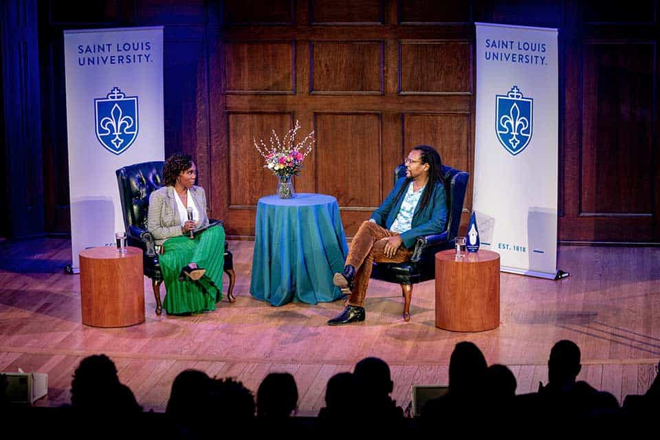 Two people sit on a stage in front of an audience. SLU banners are placed behind them.