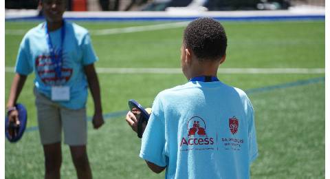 Access Academies 2024 Summer Program A student wearing an Acess Academies T-shirt is seen from behind while talking to another student on a grassy field.
