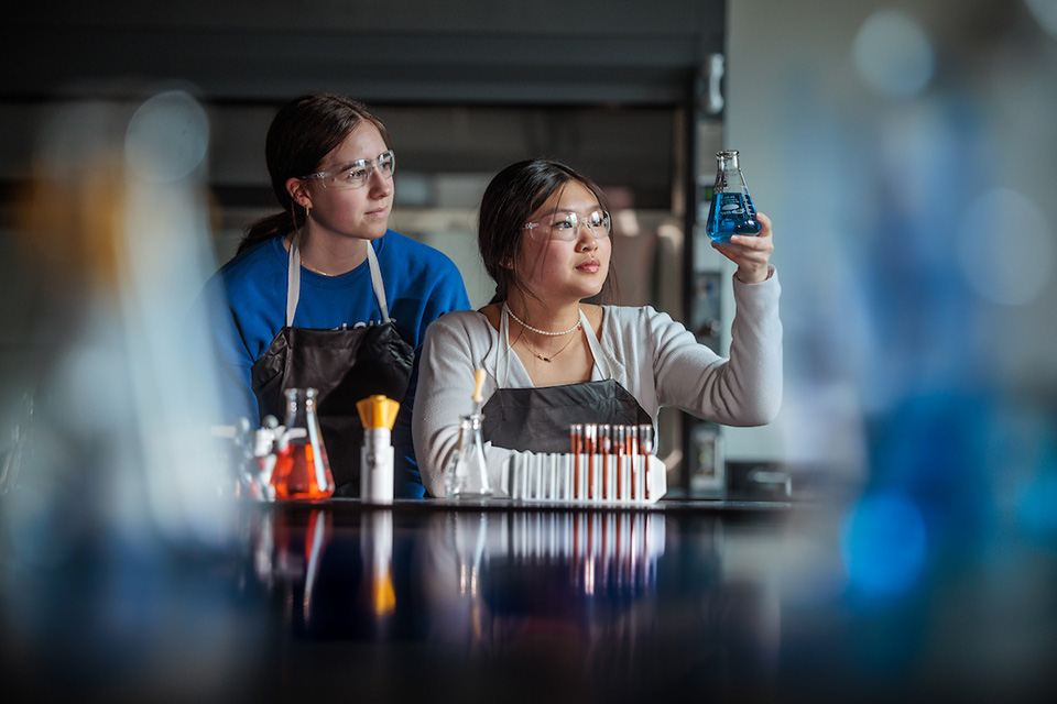 Two students work together at a lab table wearing safety googles and examining a beaker.
