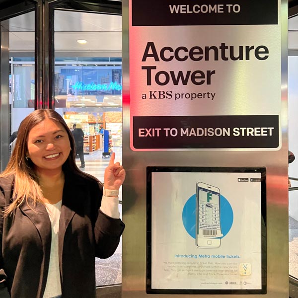 Daniella Soriano Success Story Daniella Soriano standing to the left of Accenture Tower sign. She is smiling and pointing at the sign. The full sign reads "Welcome to Accenture Tower, a KBS property. Exit to Madison Street."