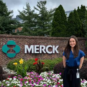 Lauren Favell Success Story Lauren Favell standing to the right of a flower bed. Above the flower bed is a large Merck logo on a brick wall.