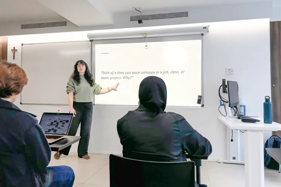 Professor stands at the front of a classroom speaking with students seated at desks. A projector screen displays "Think of a time you were unhappy in a job, class or team project. Why?