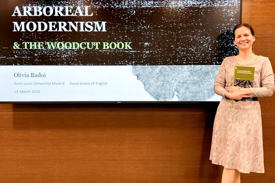 Olivia Badoi, Ph.D., poses with her book in front of the screen in San Ignacio Hall Auditorium. Woman in dress smiles at camera while holding her book in front of screen with presentation slide showing behind her.