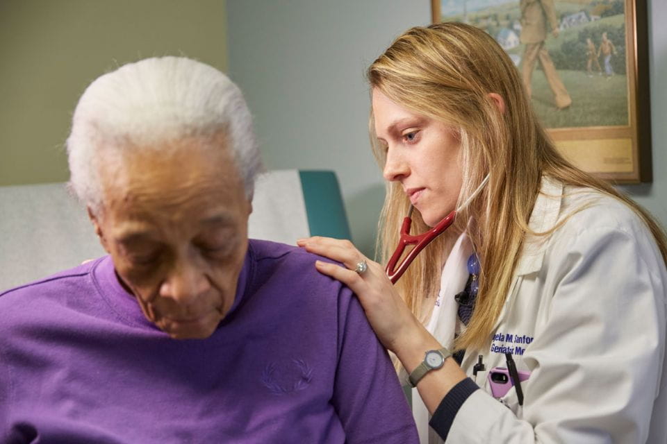 Inside a hospital exam room, a physician listens with her stethoscope on the back of an older adult patient. 