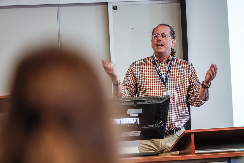 Dr. Fred Rottnek gives a lecture in a classroom, speaking animatedly with raised hands. He wears a checkered shirt and glasses, standing behind a podium.