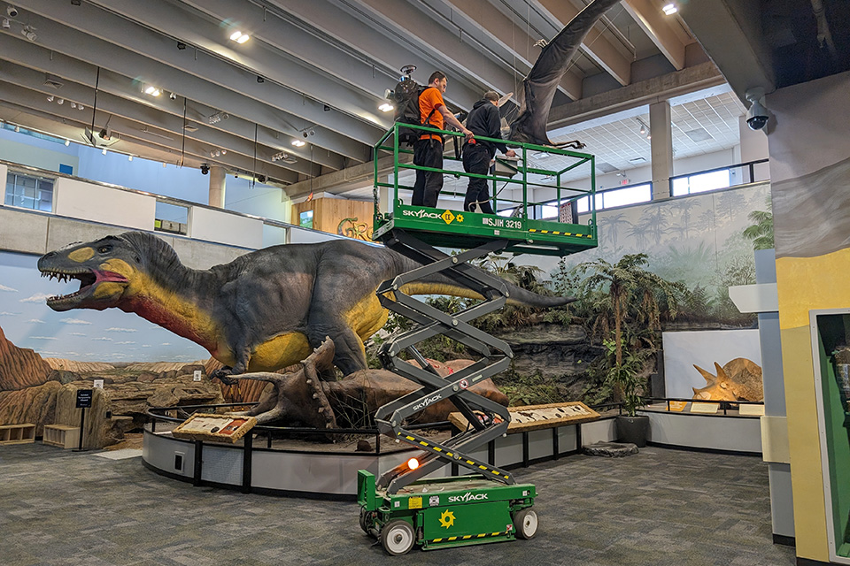 Cagri Gul, master’s student in geographic information science, stands atop the SkyJack as Chris Lucas, exhibit designer with the Saint Louis Science Center, drives. The scanning team needs to capture angles of the diorama from above in order to create a more complete scan; simply walking with the backpack on the ground would not collect data from the top of the dinosaurs. Cagri Gul, master’s student in geographic information science, stands atop the SkyJack as Chris Lucas, exhibit designer with the Saint Louis Science Center, drives. The scanning team needs to capture angles of the diorama from above in order to create a more complete scan; simply walking with the backpack on the ground would not collect data from the top of the dinosaurs.