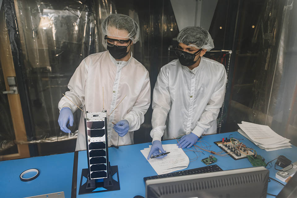 Michael Dompke (left), Senior Mechanical Engineering student and DARLA project manager working with Junior Engineering student Adrian Acevedo to prepare DARLA for launch. Two students wearing protective coats, face masks, gloves and head wear, examine a piece of equipment on a table.